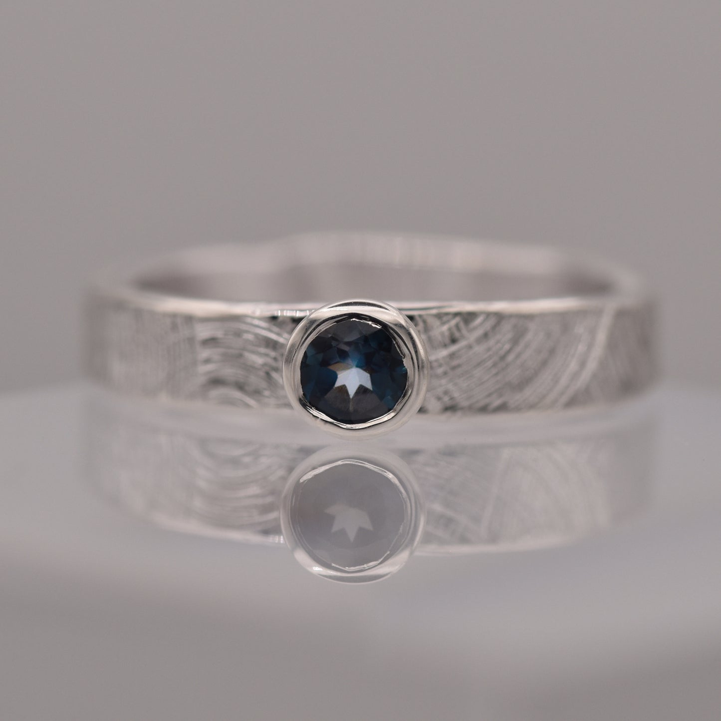 Silver (patterned with concentric circles to symbolise tree rings and growth) ring with a blue gemstone on a reflective surface