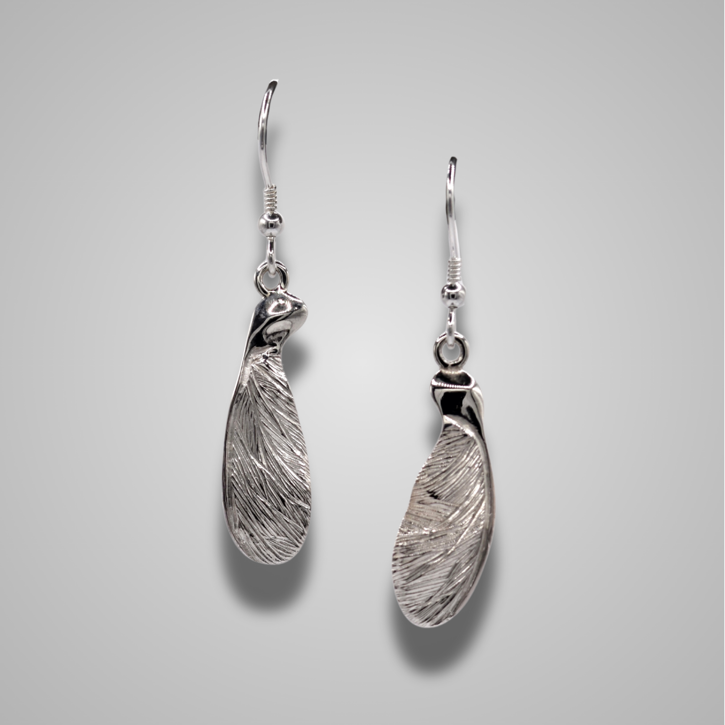 A pair of sterling silver earrings on silver hooks. The earrings are half sycamore seeds hanging down to create long drop earrings with sparkly engraved details.