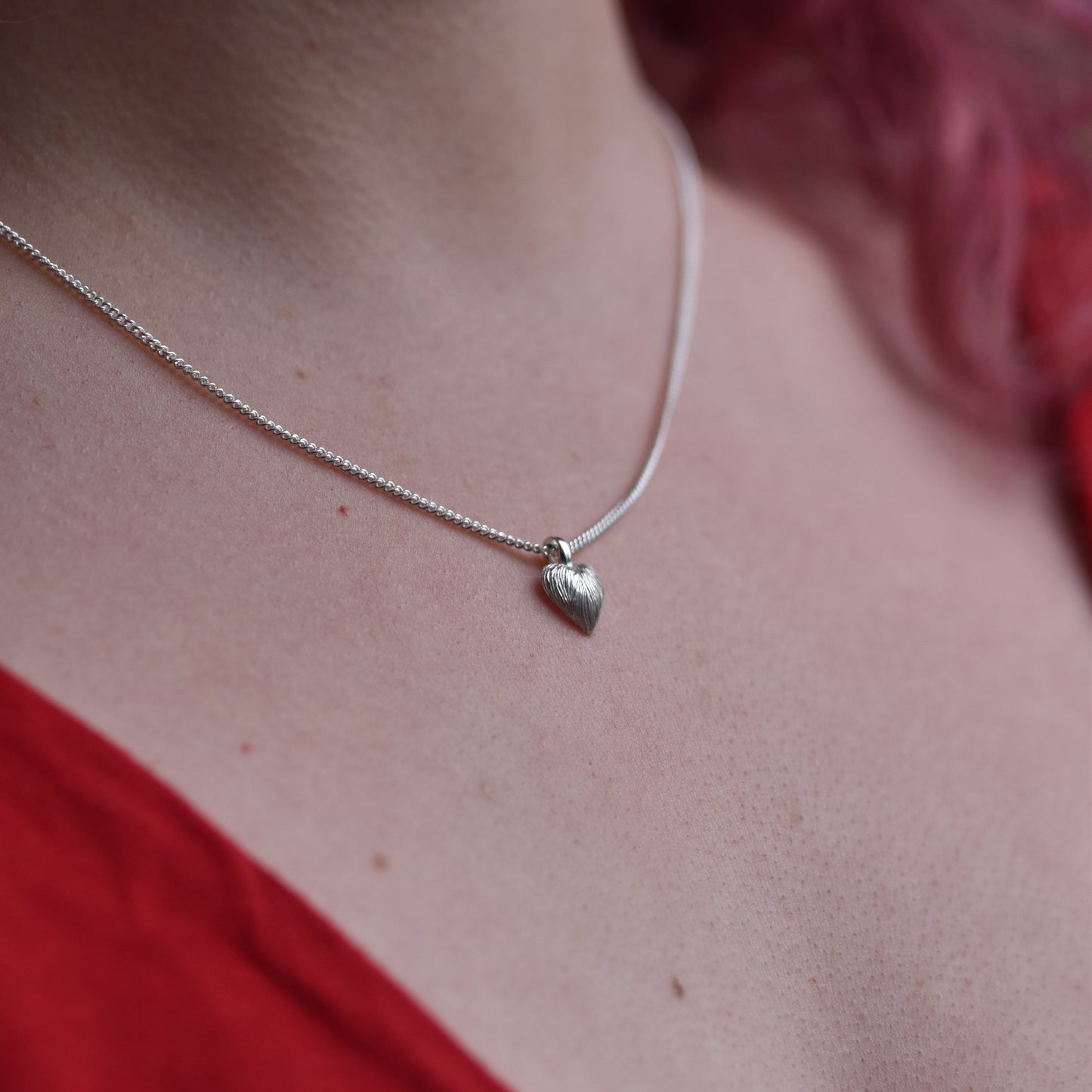 Girl with red hair and a red top is wearing a small delicate heart pendant. The pendant is made of silver with sparkly engraved lines on it.
