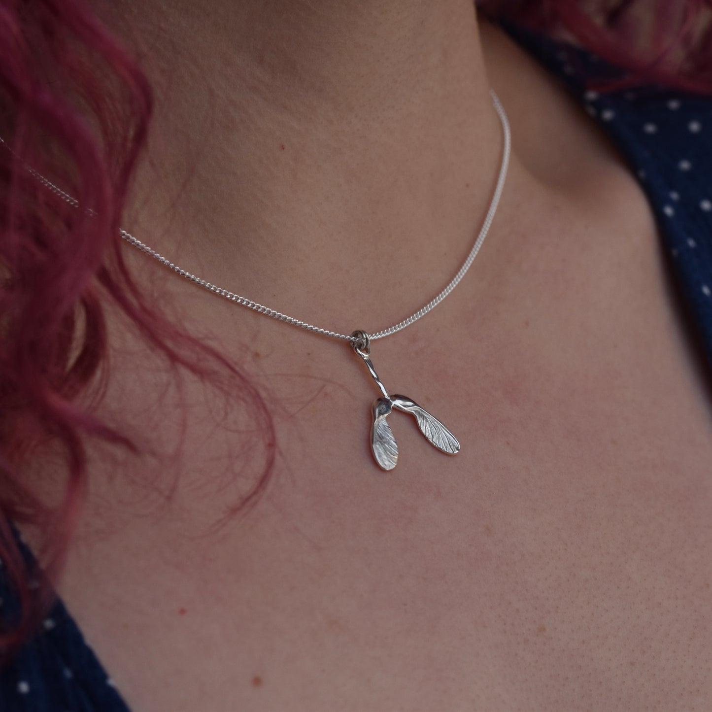Girl wearing a blue dress, with red hair is wearing a sycamore seed pendant that is embellished with engraving and highly polished silver