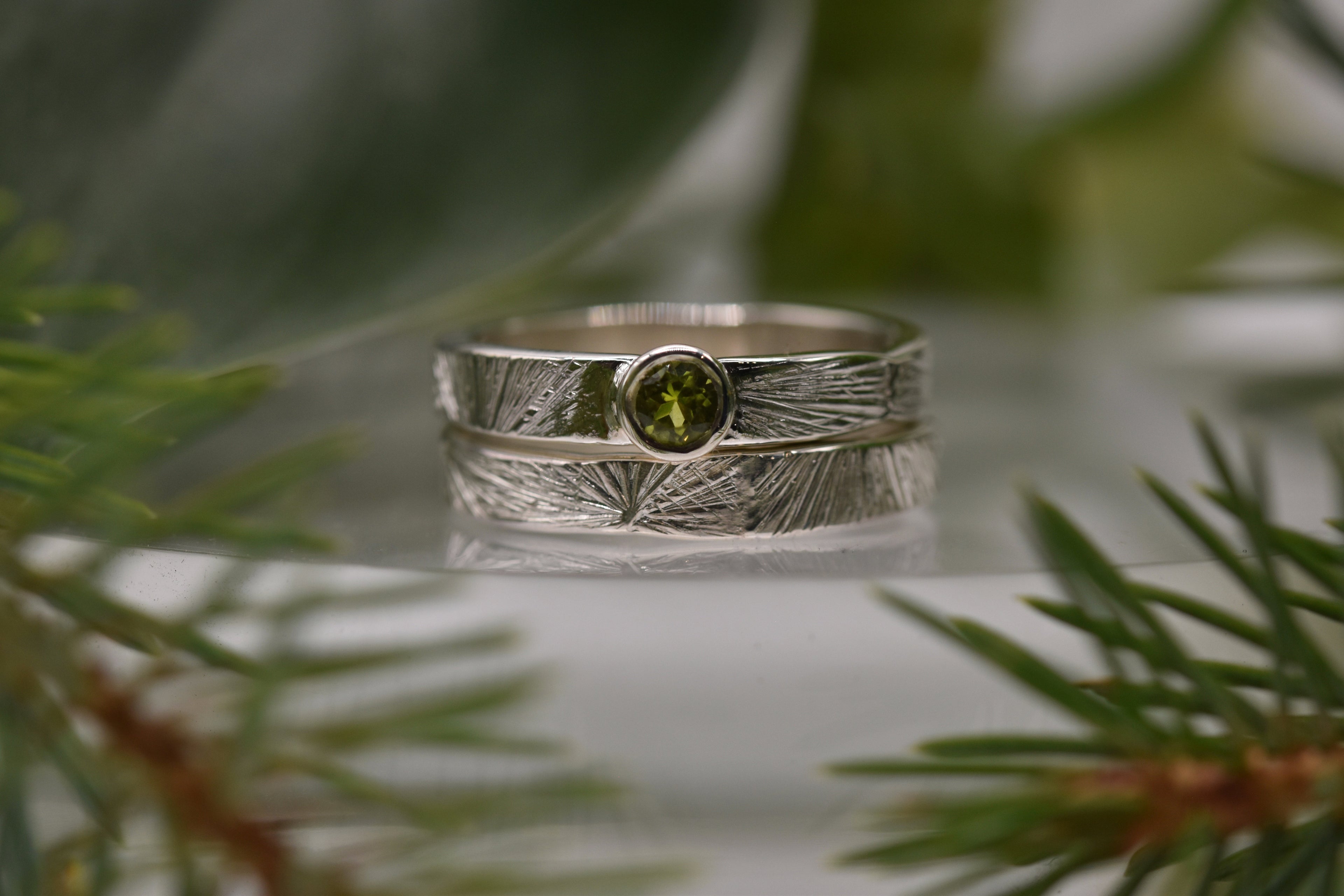 green gemstone set in a silver ring with unusual radiating pattern. stacked ontop of a plain ring with the same pattern. The picture is framed with green pine needles