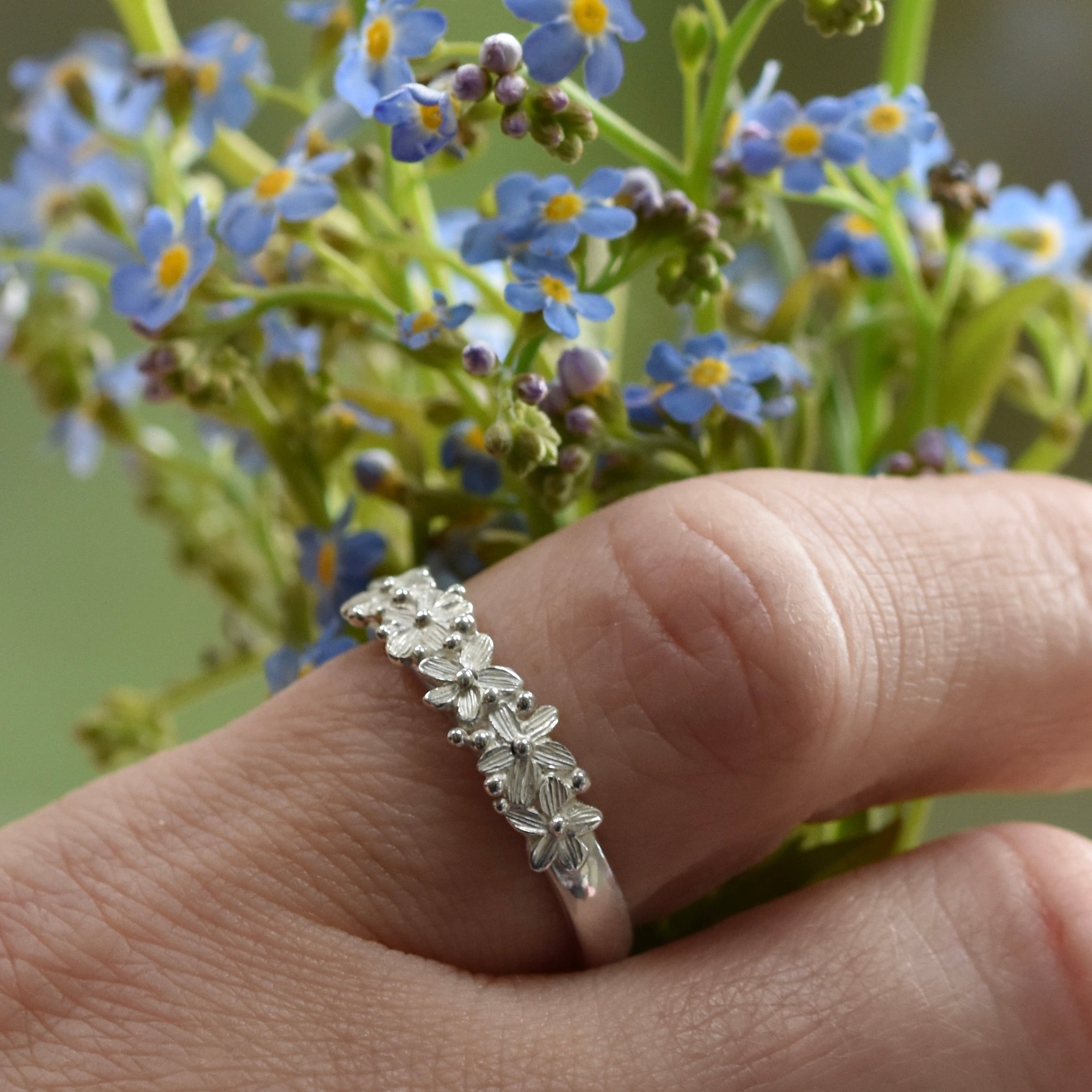hand wearing a silver ring with forget me not flowers accross the front of the band. The hand is also holding a bunch of blue forget me not flowers
