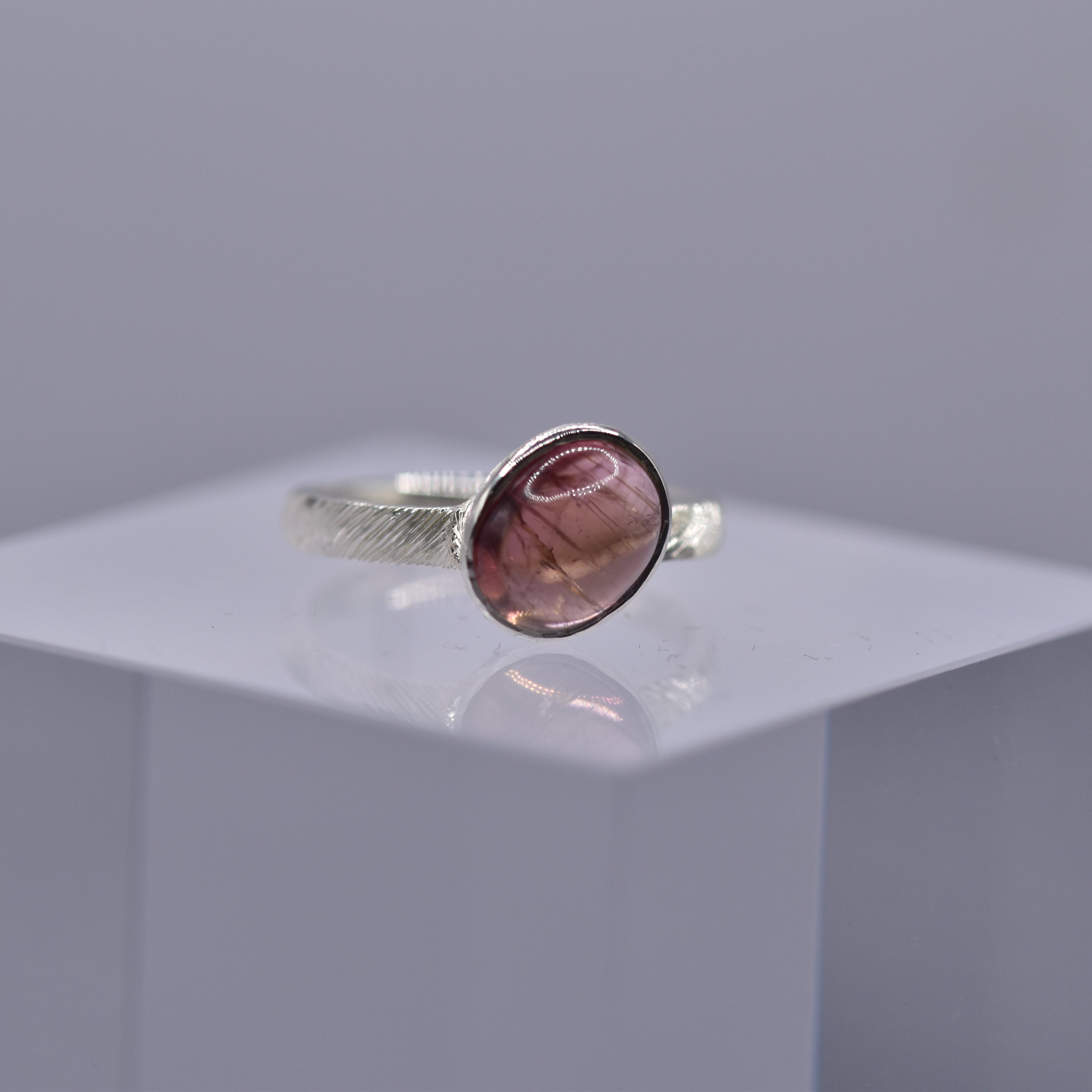 Silver ring with a pinkish-purple stone on a clear cube with a gray background