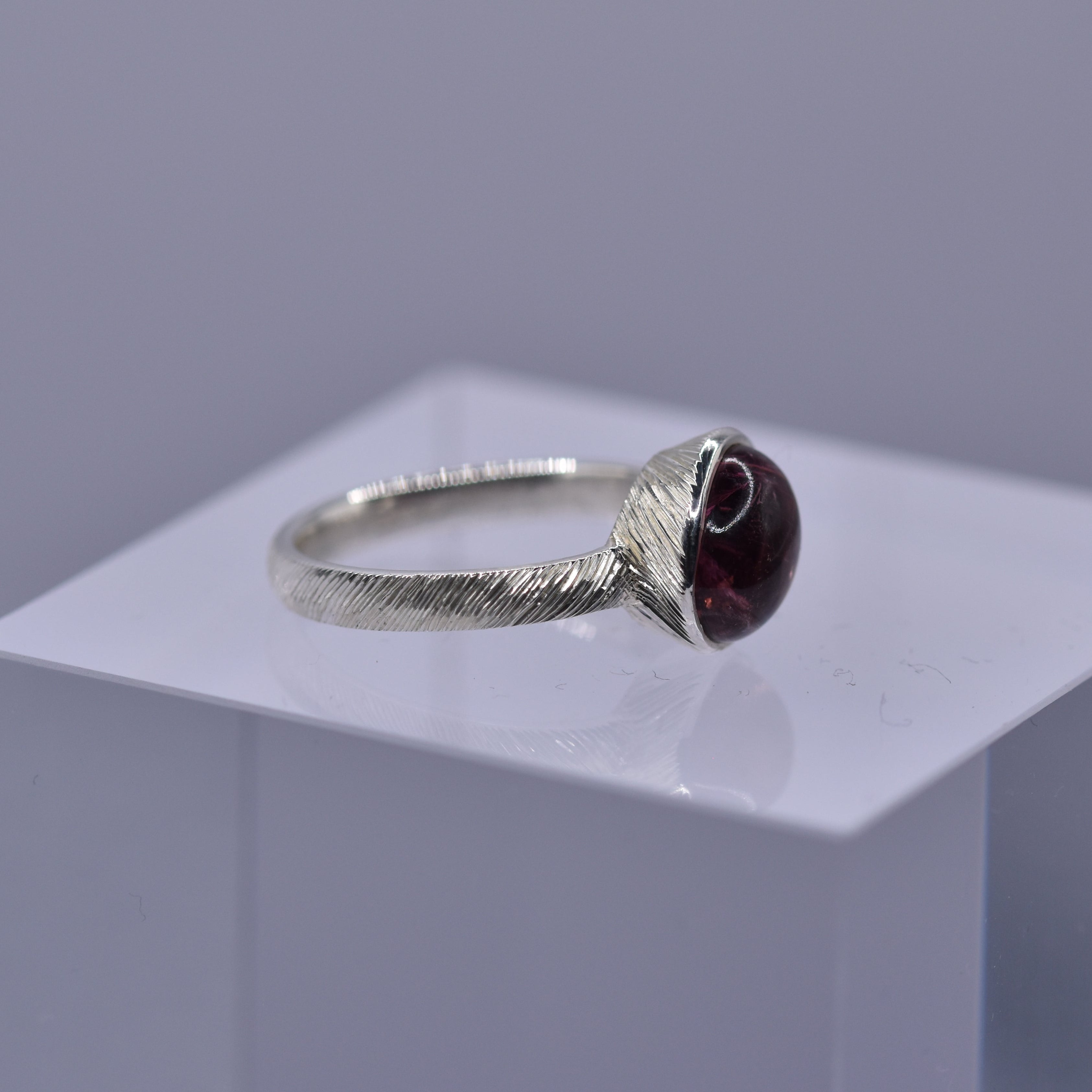 Silver ring with a pinkish gemstone with textured hand engraved lines on a clear cube against a gray background