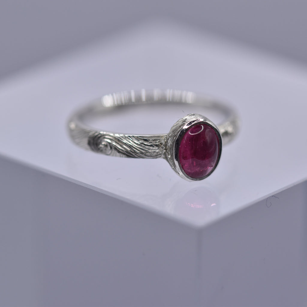 Silver ring with a pink gemstone with swirls and waves engraved on the ring on a gray background