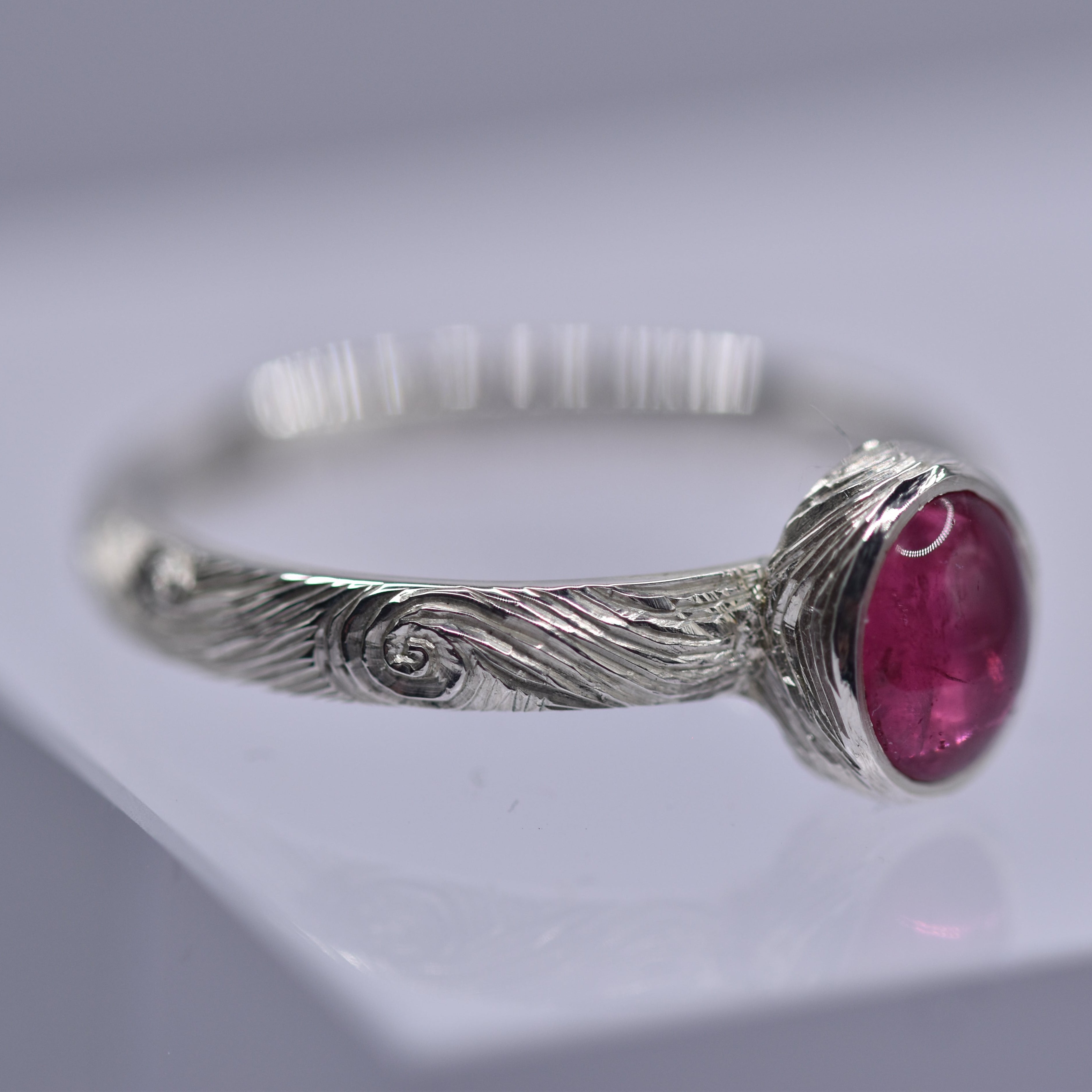 Silver ring with a pink gemstone with swirls and waves engraved on the ring on a gray background
