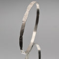 A polished silver bangle bracelet with a textured, sunburst-like engraved pattern on its exterior, displayed on a neutral gray reflective surface.