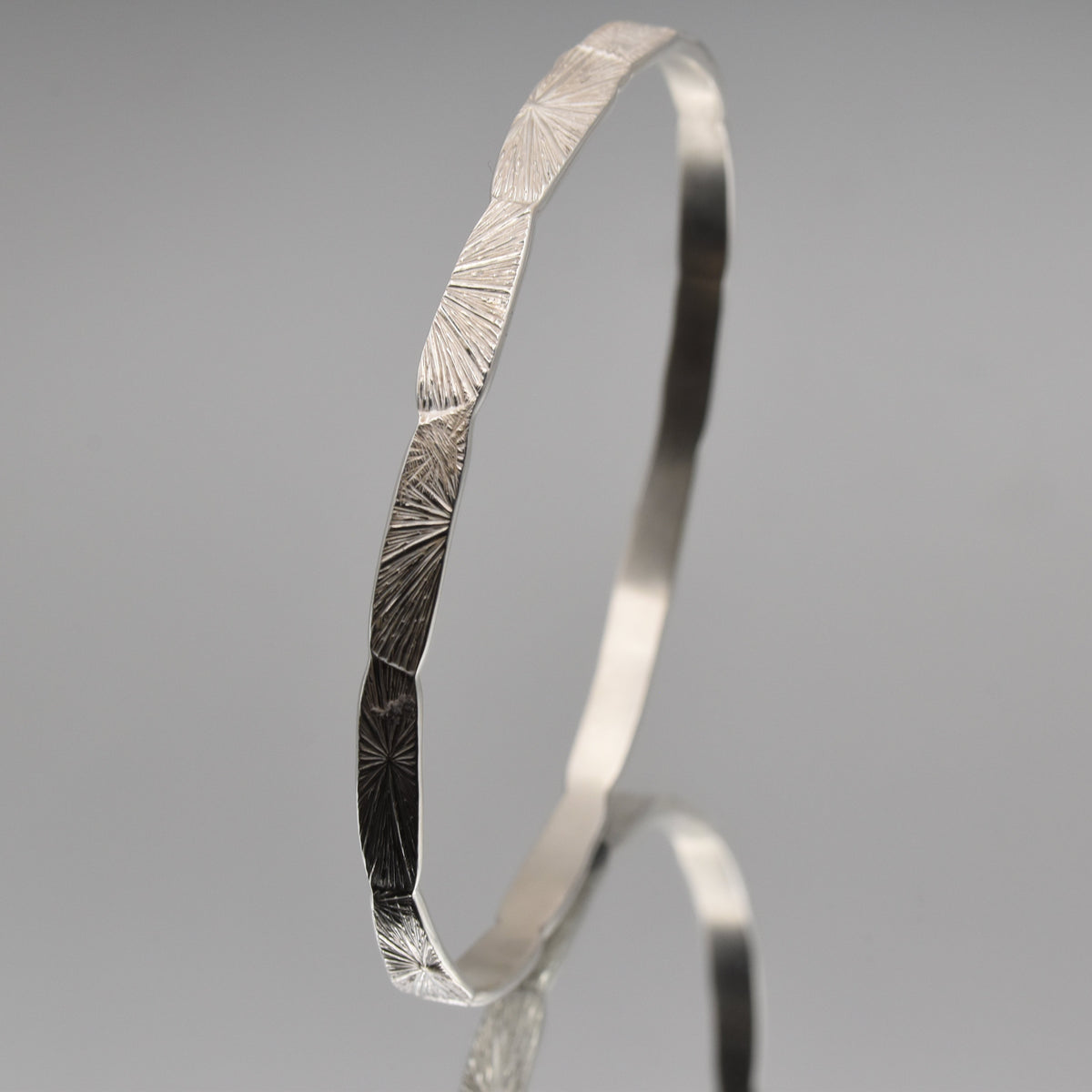 A polished silver bangle bracelet with a textured, sunburst-like engraved pattern on its exterior, displayed on a neutral gray reflective surface.