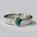 A sterling silver ring with a textured band and a central bezel-set round turquoise gemstone, shown on a neutral white background.