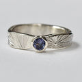 A silver ring with a textured sunburst pattern on the band and a round lilac gemstone in the centre, photographed on a white background.