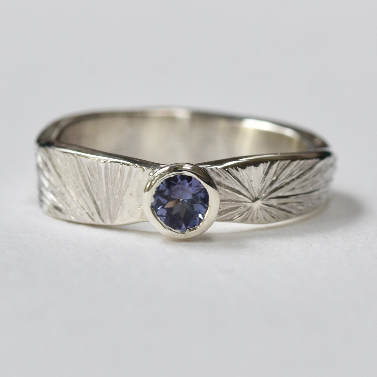 A silver ring with a textured sunburst pattern on the band and a round lilac gemstone in the centre, photographed on a white background.