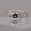 Silver (patterned with concentric circles to symbolise tree rings and growth) ring with a blue gemstone on a reflective surface
