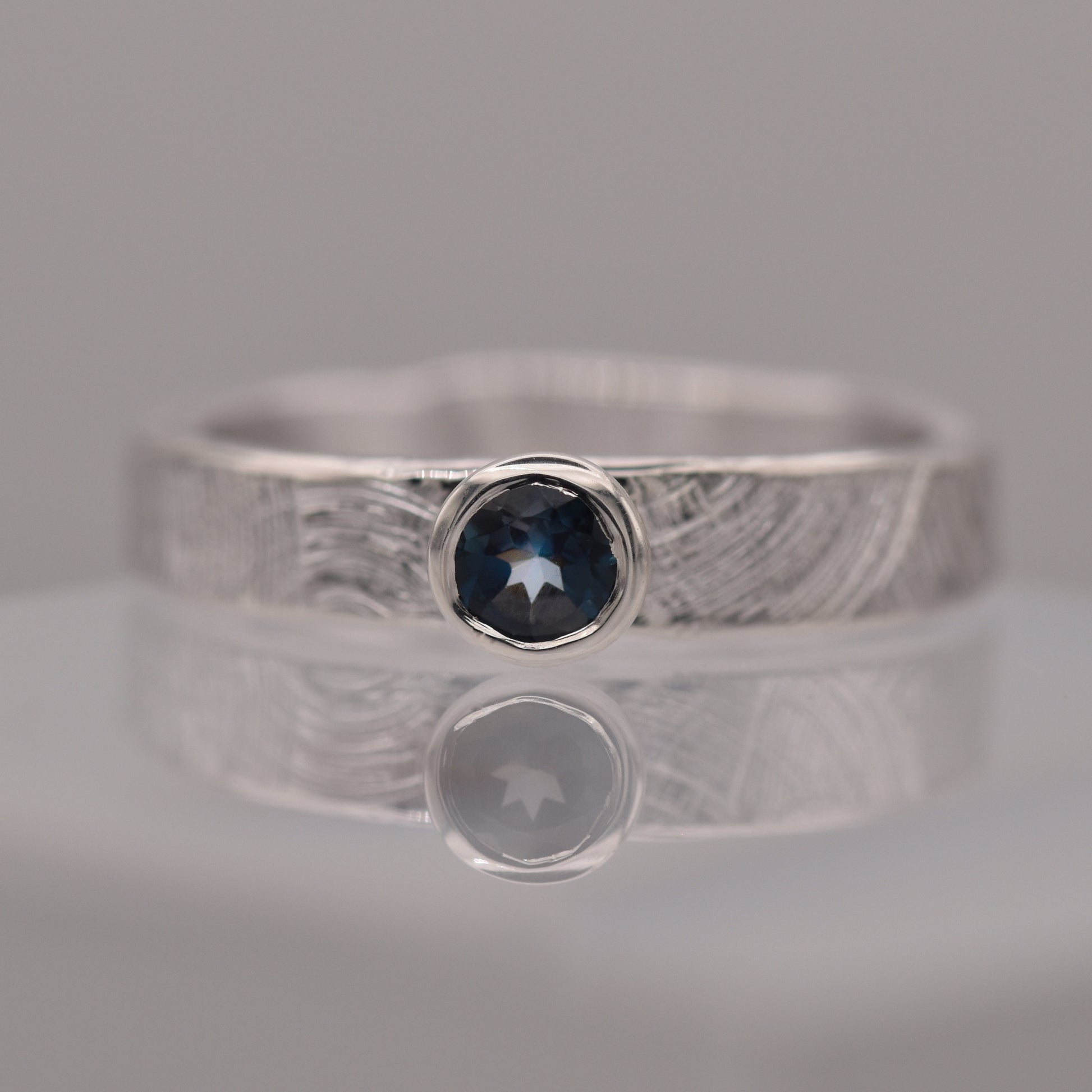 Silver (patterned with concentric circles to symbolise tree rings and growth) ring with a blue gemstone on a reflective surface