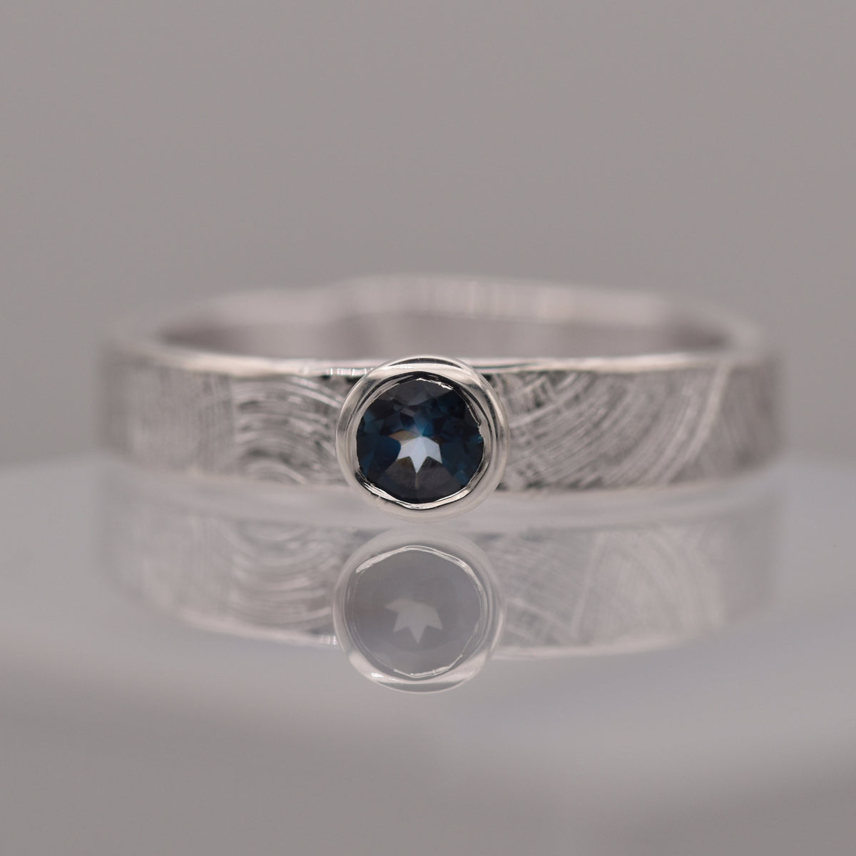 Silver (patterned with concentric circles to symbolise tree rings and growth) ring with a blue gemstone on a reflective surface