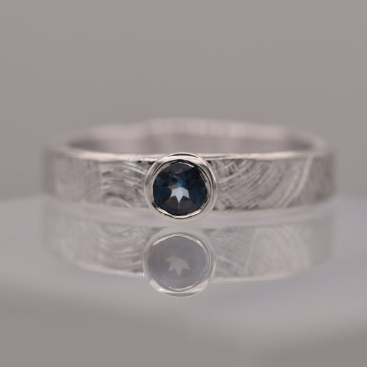 Silver (patterned with concentric circles to symbolise tree rings and growth) ring with a blue gemstone on a reflective surface