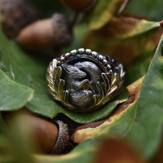 Silver ring with sleeping squirrel clutching an orange sapphire and nestled among oak leaves with a nature-themed design on green leaves and acorns.