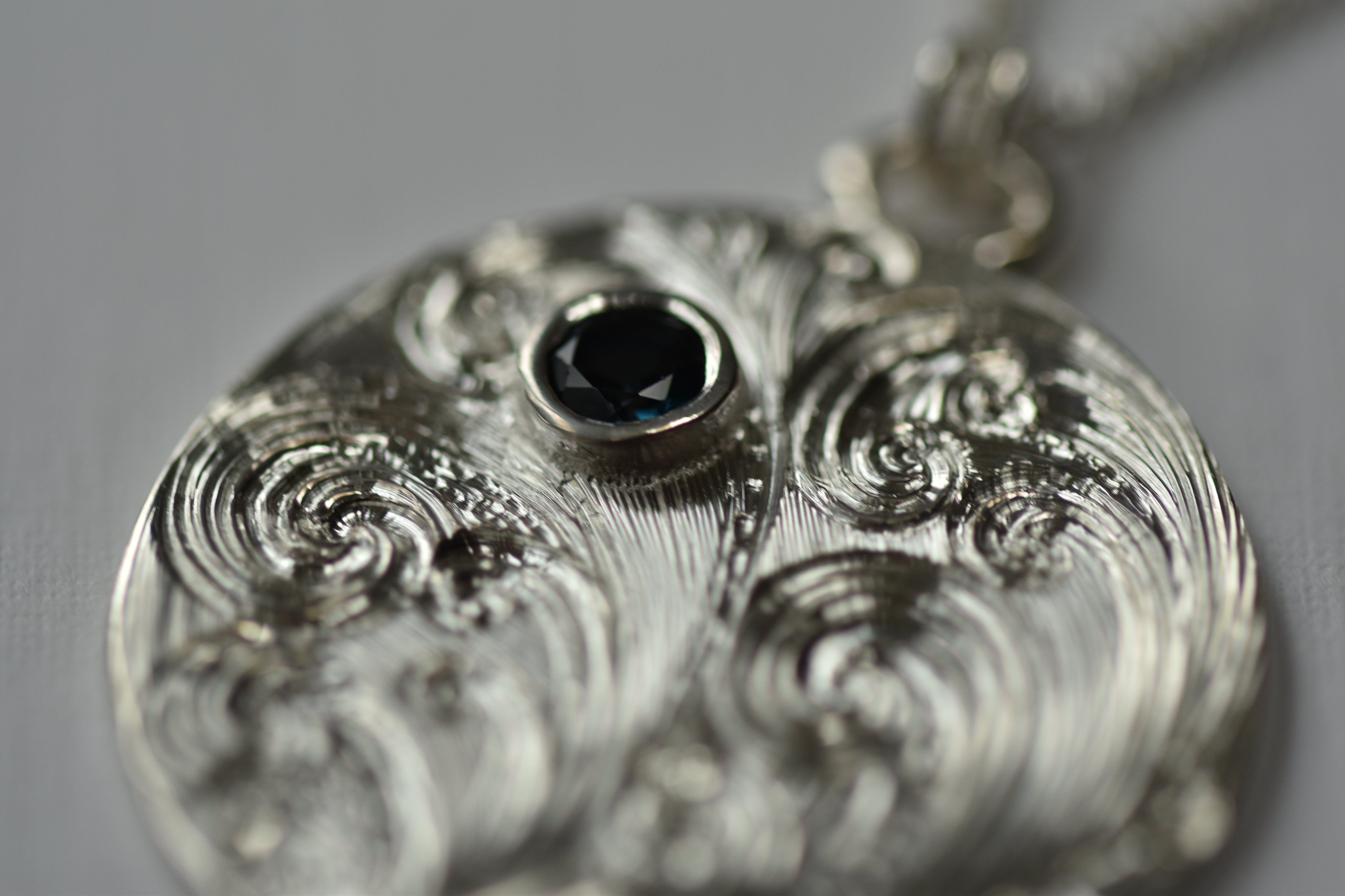 Silver pendant with intricate design and central blue gemstone on a gray background