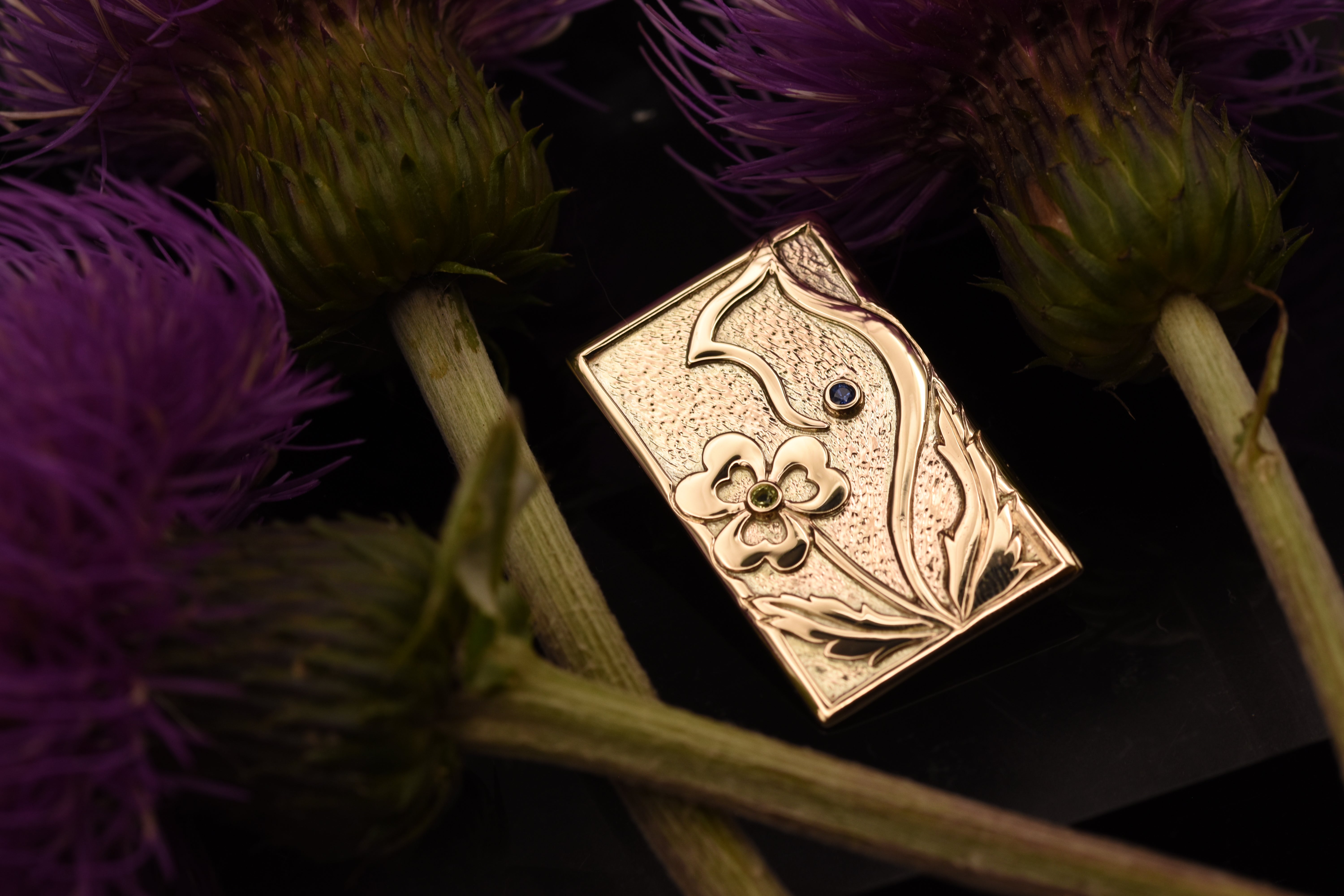 A rectangular gold brooch with thistle and shamrock motifs and a small blue and green gemstones, photographed among purple thistle flowers on a dark background.