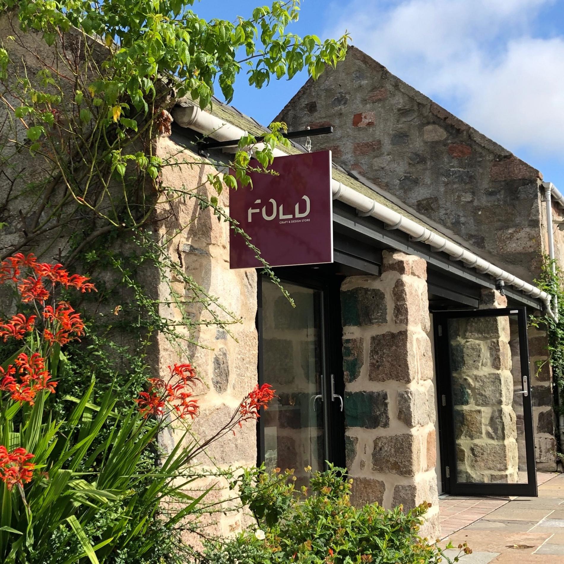 Stone building with a 'FOLD' sign, surrounded by plants and flowers