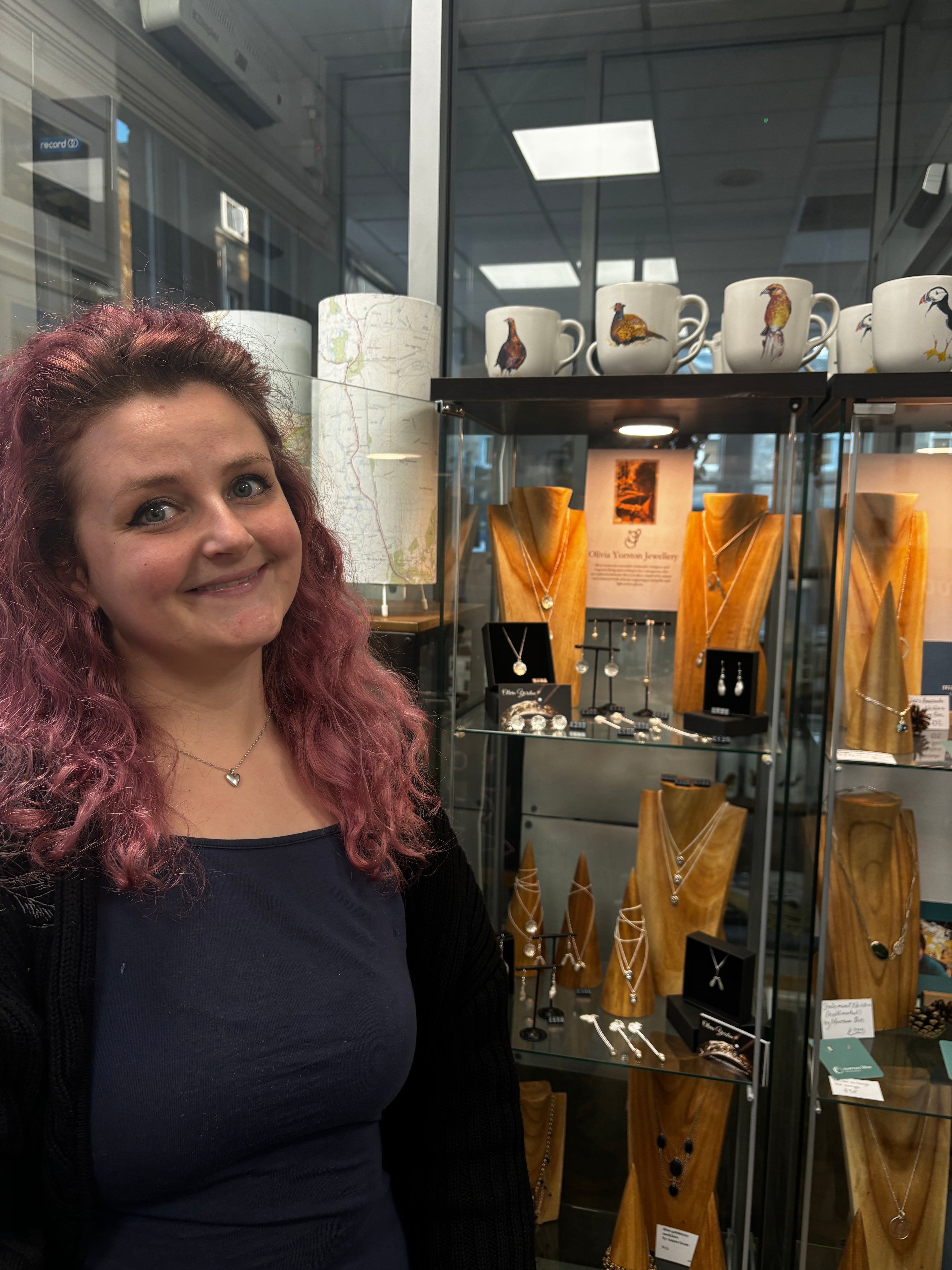Woman with pink hair standing in front of a display case with jewelry and mugs.
