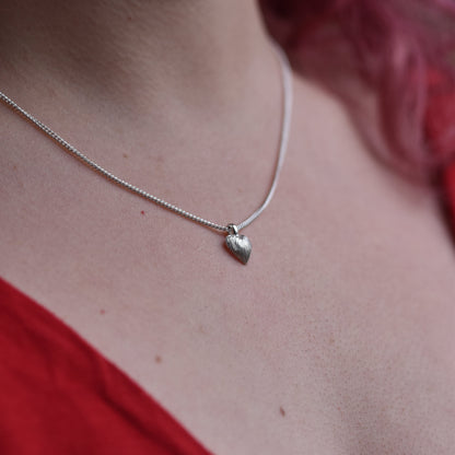 Girl with red hair and a red top is wearing a small delicate heart pendant. The pendant is made of silver with sparkly engraved lines on it. 