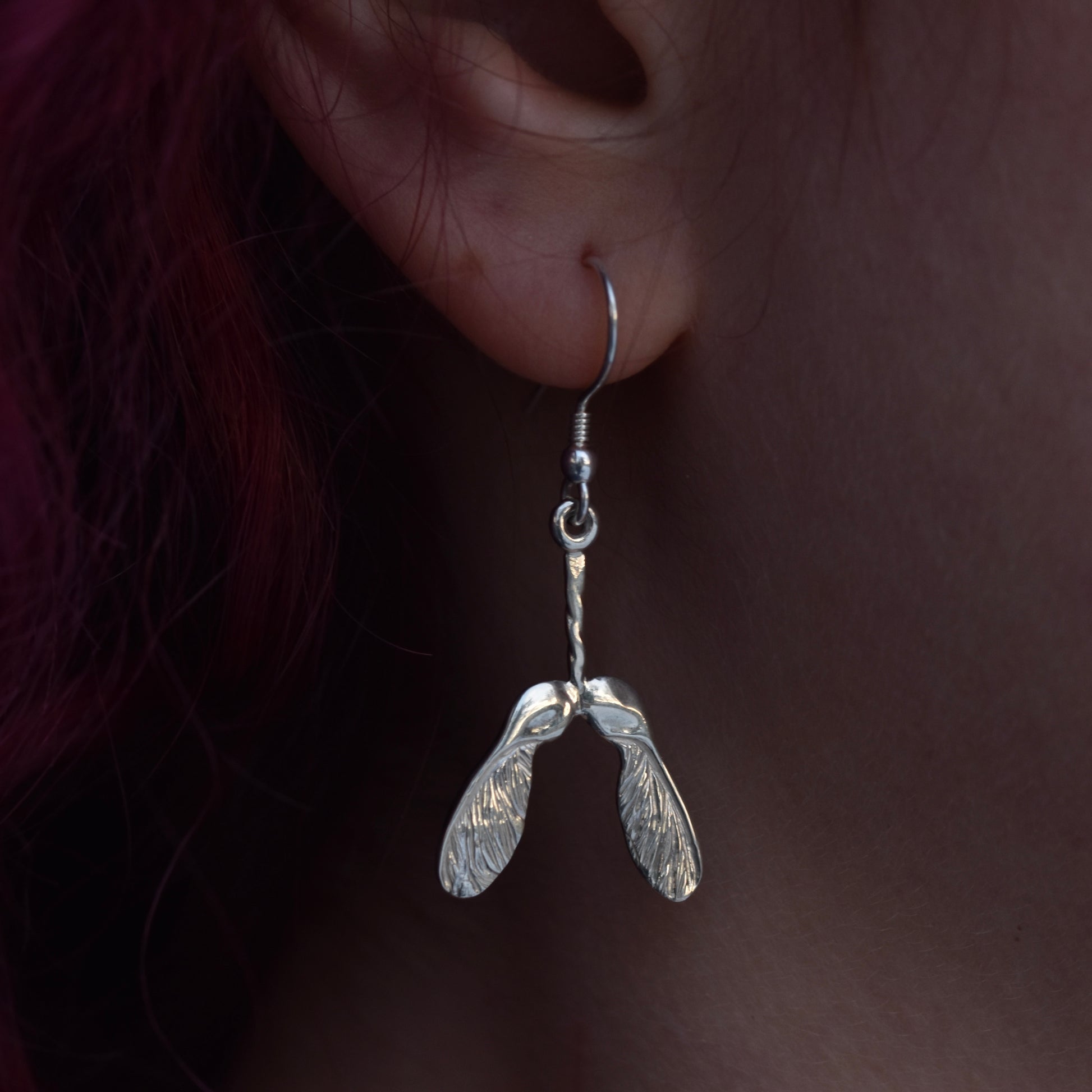 Very shiny earring worn by a girl with red hair. The earring is a sycamore seed covered in sparkley engraving with highly polished silver on a silver hook 