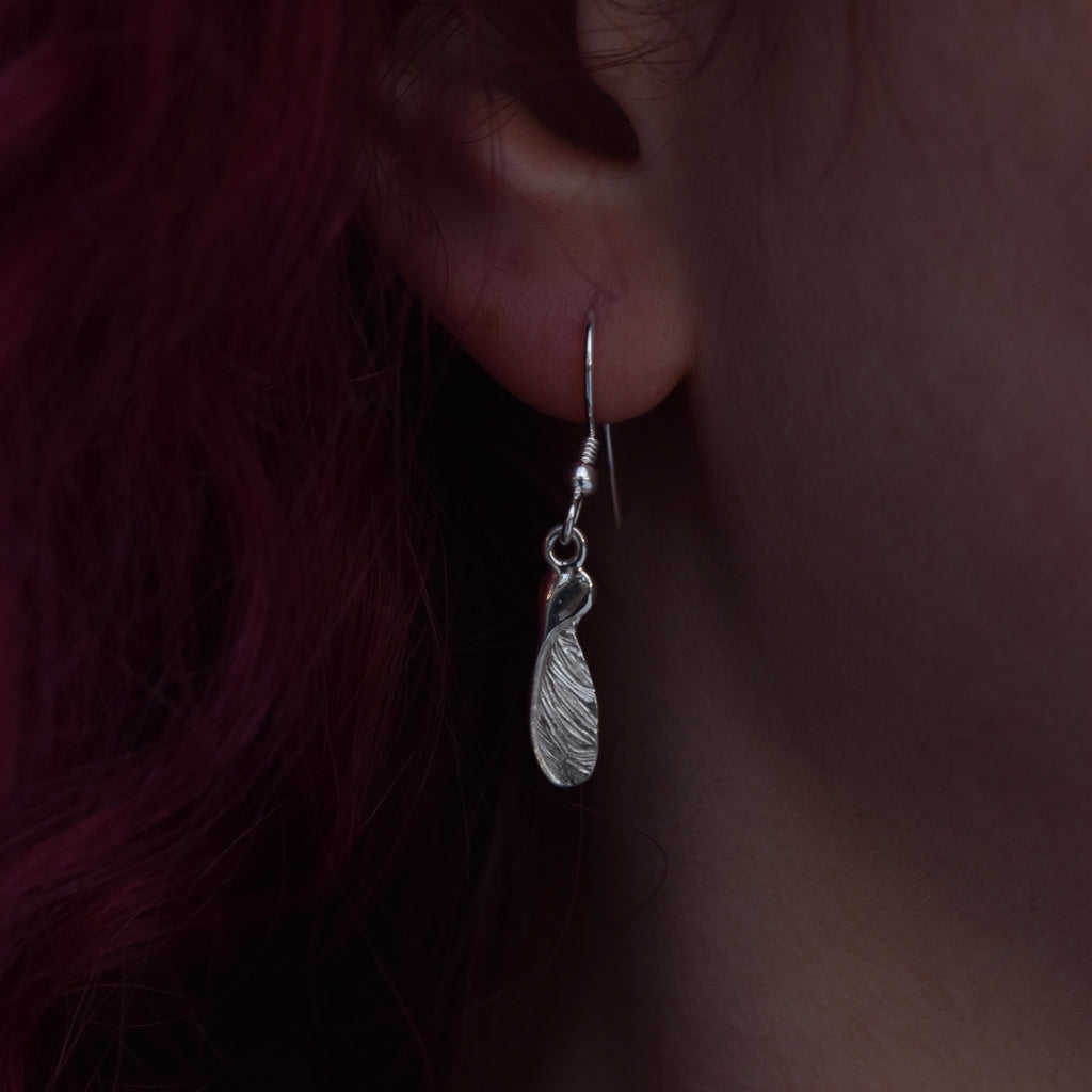 Girl with red hair wearing a medium drop earring representing a highly detailed and realistic half sycamore seed earring made from silver on a silver hook