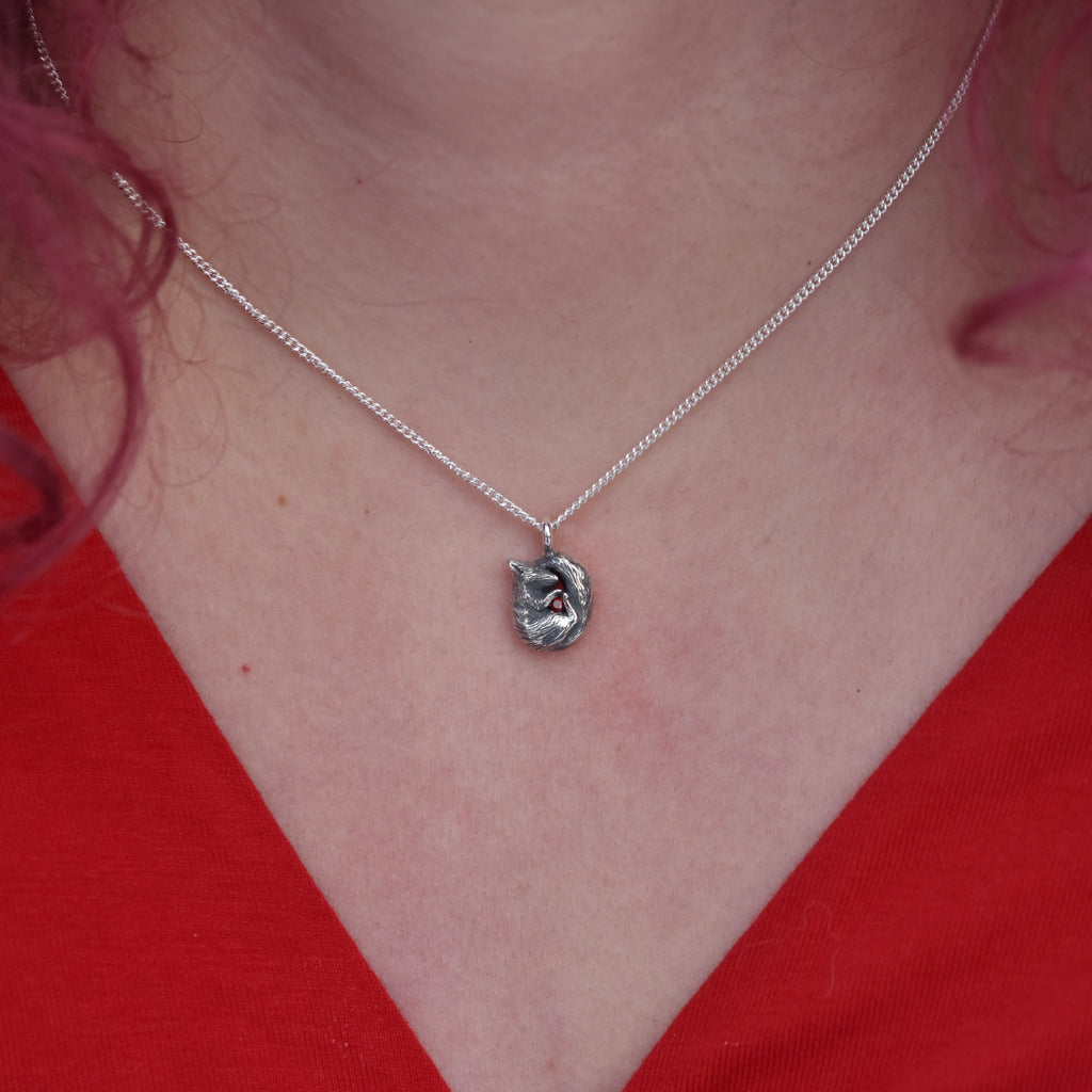 Girl with red hair and v necked top on is wearing a pendant made of silver. The pendant is a tiny curled up squirrel clutching a red gemstone which is a garnet 